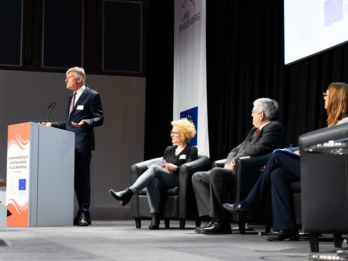 Bild: Am Rednerpult auf der Bühne steht Egbert Holthuis, Vertreter der EU-Kommission. Auf schwarzen Sesseln sitzen die Moderatorin, Wirtschaftsminister Steinbach und EU-Kommissionsvertreterin Zademach-Schwierz.