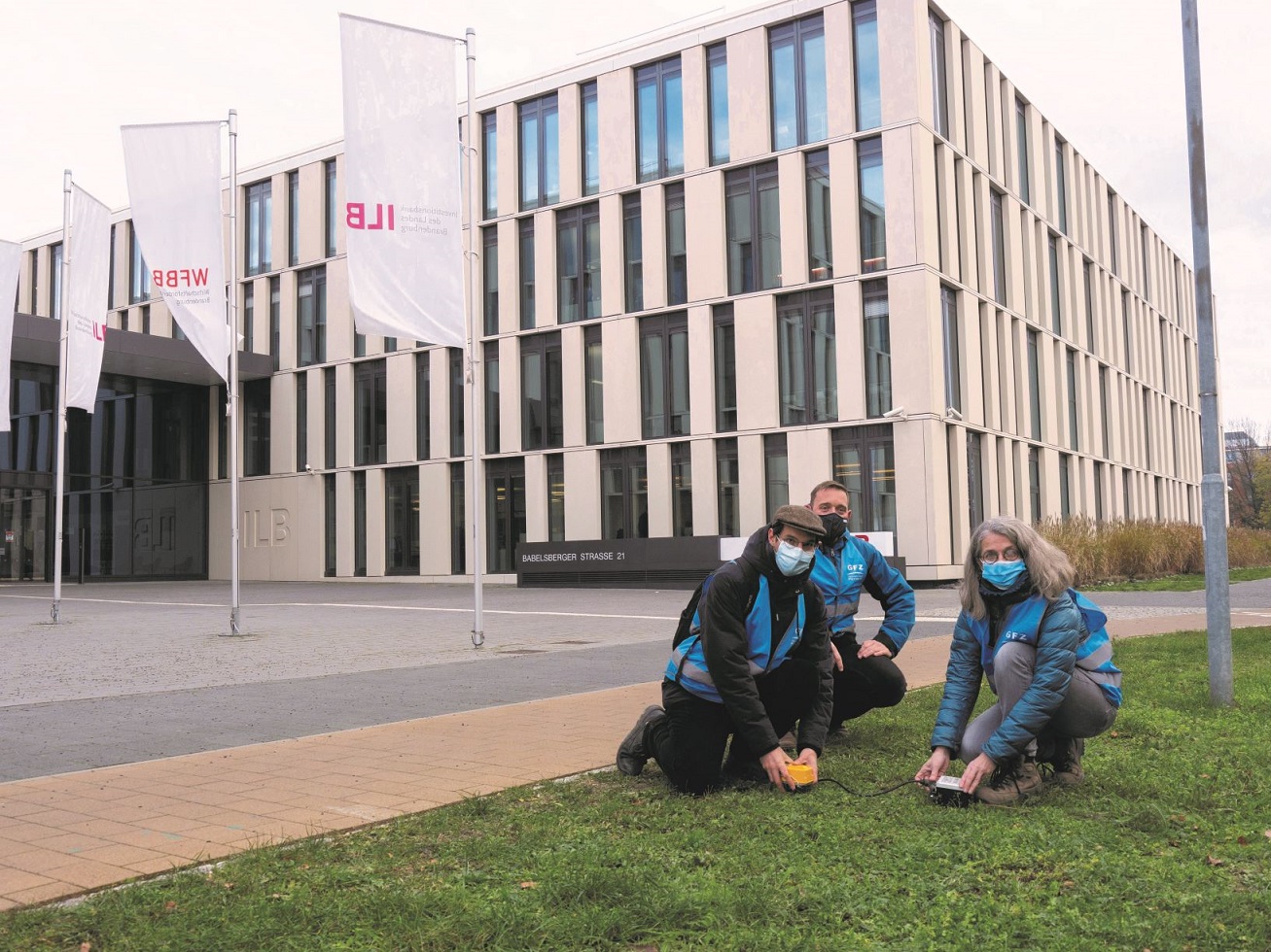 Vor dem Gebäude der Investitionsbank des Landes Brandenburg in Potsdam hocken drei Forschende auf dem begrünten Boden mit Messgeräten in der Hand.