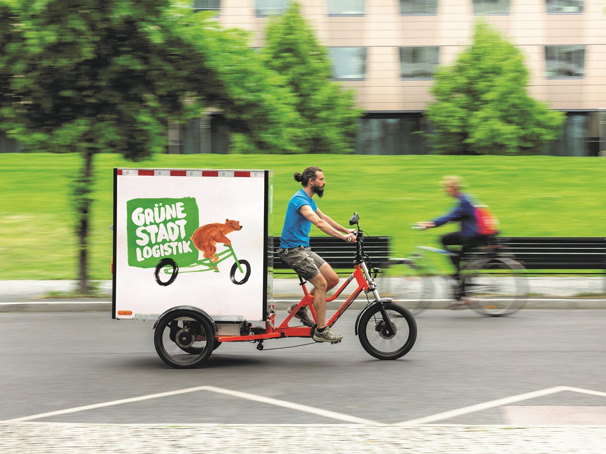 Ein Mann fährt auf einem Lastenrad durch eine Straße. Auf dem Anhänger des Lastenrads ist ein großer Kasten mit der Aufschrift Grüne Stadtlogistik zu sehen. 