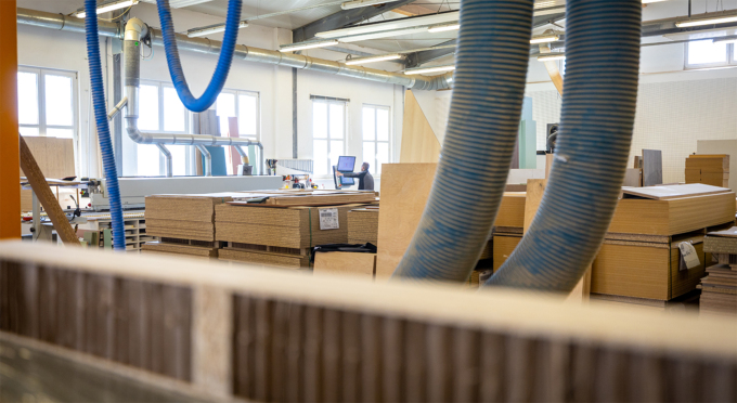 Innenansicht der Tischlerei mit großen Stapeln von Holzplatten und mehreren großen flexiblen Rohren, die von der Decke hängen