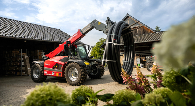 Ein roter Teleskoplader hebt eine große Rolle aus schwarzem Rohrmaterial an. Im Hintergrund sind Lagergebäude und vorne Pflanzen sichtbar.