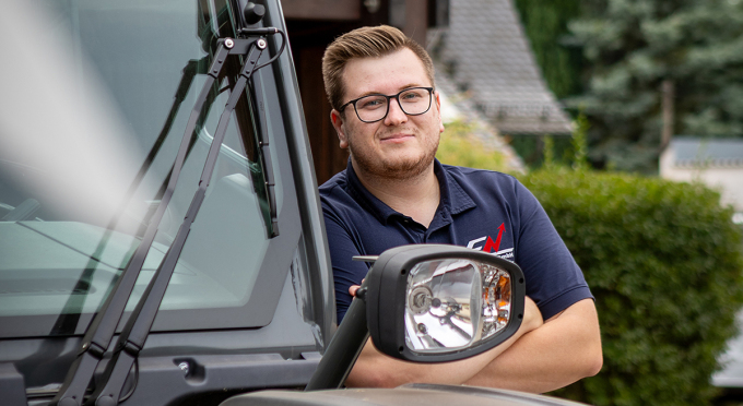 Ein Mitarbeiter der Elektro Nimtz GmbH lehnt an einem Fahrzeug. Er trägt ein dunkelblaues Polohemd mit dem Logo des Unternehmens. Der Mann trägt eine Brille und hat die Arme verschränkt.