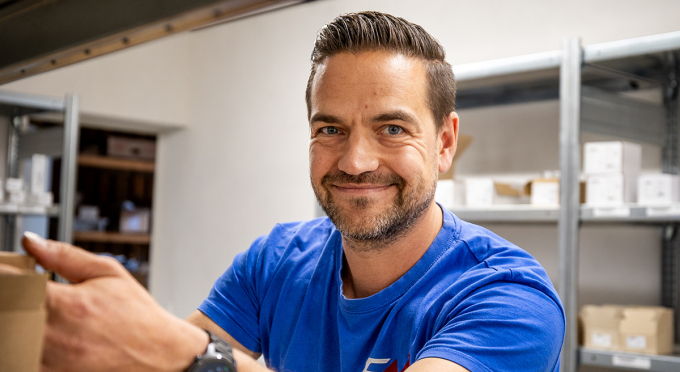 Ein Mann in einem blauen T-Shirt hält einen Karton in beiden Händen. Im Hintergrund sind Regale mit mehreren Kisten und Verpackungen zu sehen.