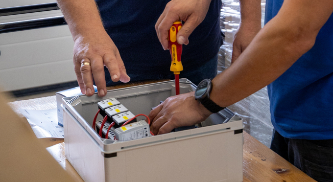 Zwei Hände arbeiten an einem offenen Gehäuse mit mehreren elektrischen Bauteilen. Eine Hand hält einen Schraubendreher, die andere Hand greift nach einem Bauteil.