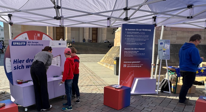 EFRE/JTF-Projektrad sowie eine Infostele mit zwei interessierten Jungen, einer Frau und einem abseits stehenden Mann beim Potsdamer Europafest 2025 