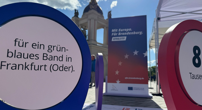 Nahaufnahme eines Standortmarkers am EFRE/JTF-Infostand mit der Aufschrift "für ein grün-blaues Band in Frankfurt (Oder)." und einer Info-Stele mit der Aufschrift "Mit Europa. Für Brandenburg. EFRE/JTF"