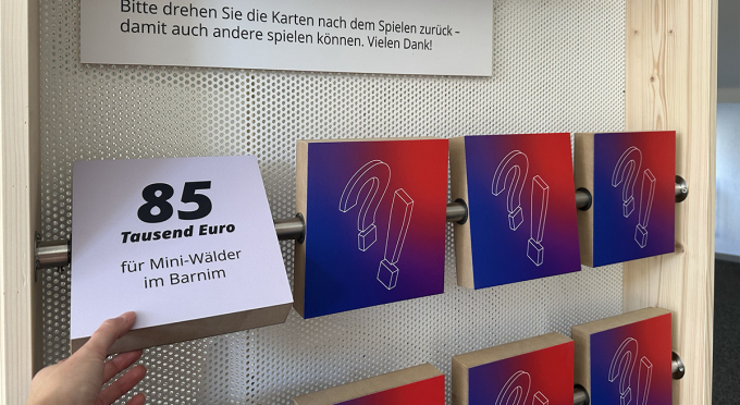 Blick auf das Holz-Memory-Spiel mit Drehmechanismus an einer Tafel der EFRE/JTF-Wanderausstellung, bei welchem das Element mit dem Schriftzug "85 Tausend Euro für Mini-Wälder im Barnim" umgedreht/sichtbar ist