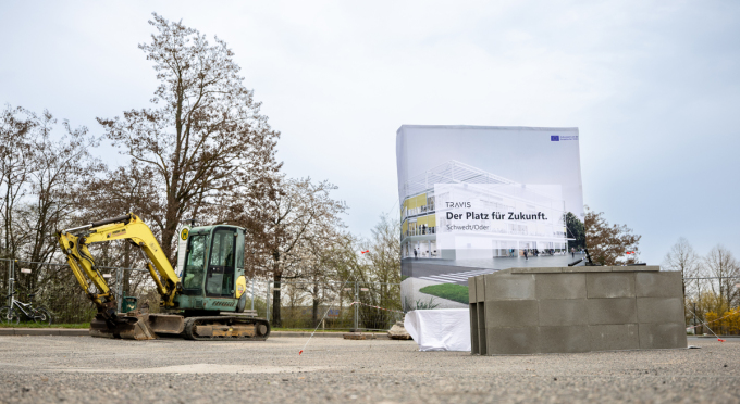 Kleiner gelber Bagger neben einem Bauzaun mit einem Banner, auf dem "TRAVIS Der Platz für Zukunft. Schwedt/Oder" steht, vor kahlen Bäumen und bewölktem Himmel