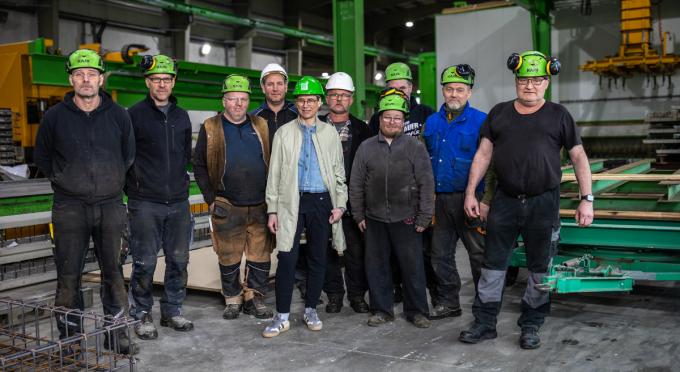 Gruppe von neun Personen der MATTIG & LINDNER GmbH in Arbeitskleidung und Schutzhelmen steht in einer Industriehalle mit Metallregalen und Maschinen im Hintergrund