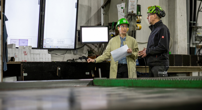 Zwei Personen mit Schutzhelmen stehen vor mehreren Bildschirmen in einer Industrieumgebung, die weibliche Person hält ein Blatt Papier